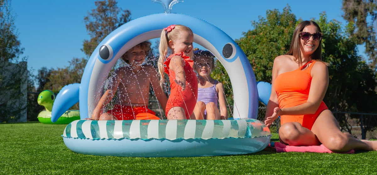 Children playing in a large inflatable Funsicle splash pool on a backyard lawn, with a woman in an orange swimsuit sitting nearby