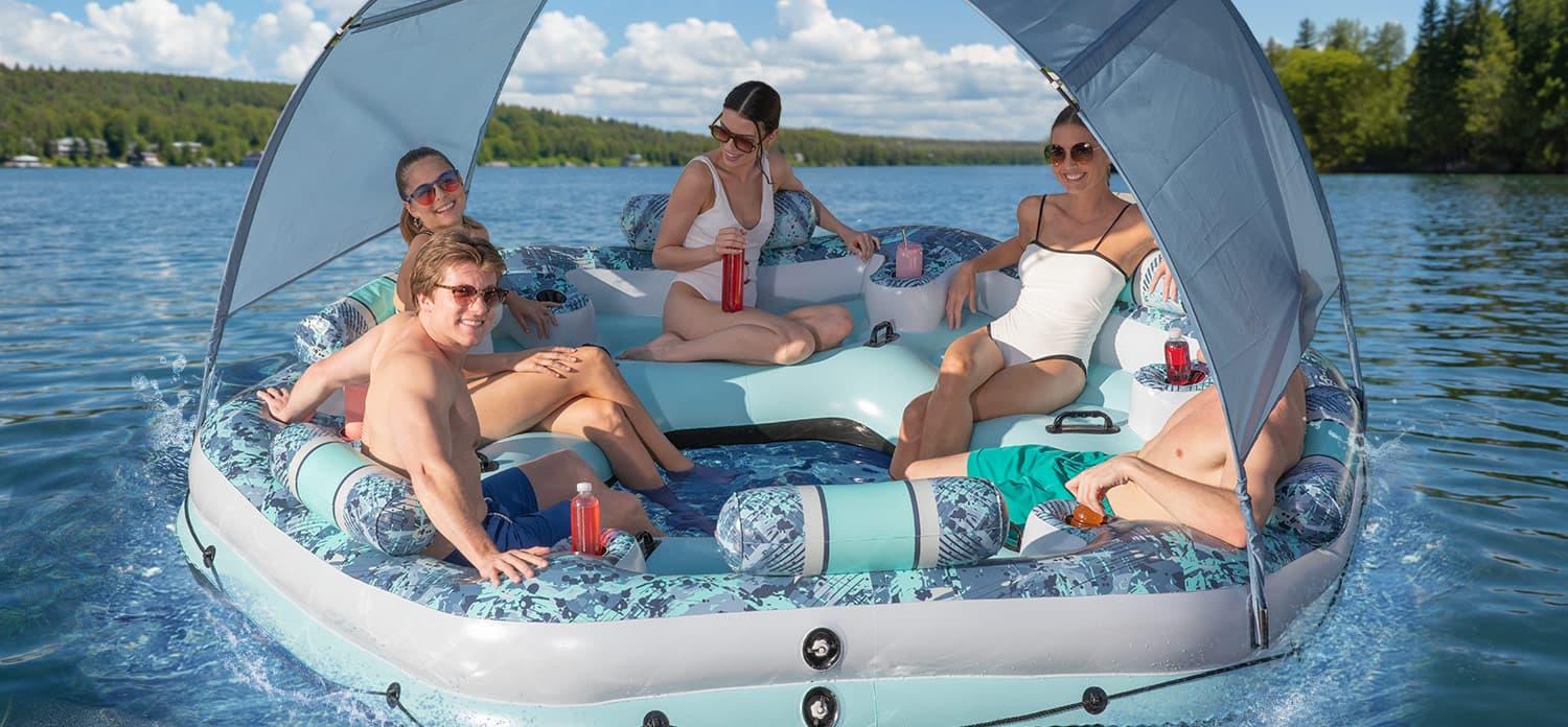 Five adults relax on a Funsicle inflatable floating lounge with a shade canopy on a sunny lake.