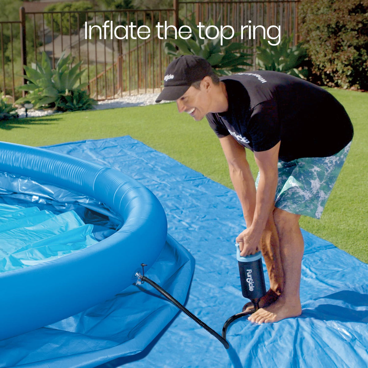 A man uses a Funsicle hand pump to inflate the blue top ring of a QuickSet Pool laid out on a protective tarp in a grassy backyard.