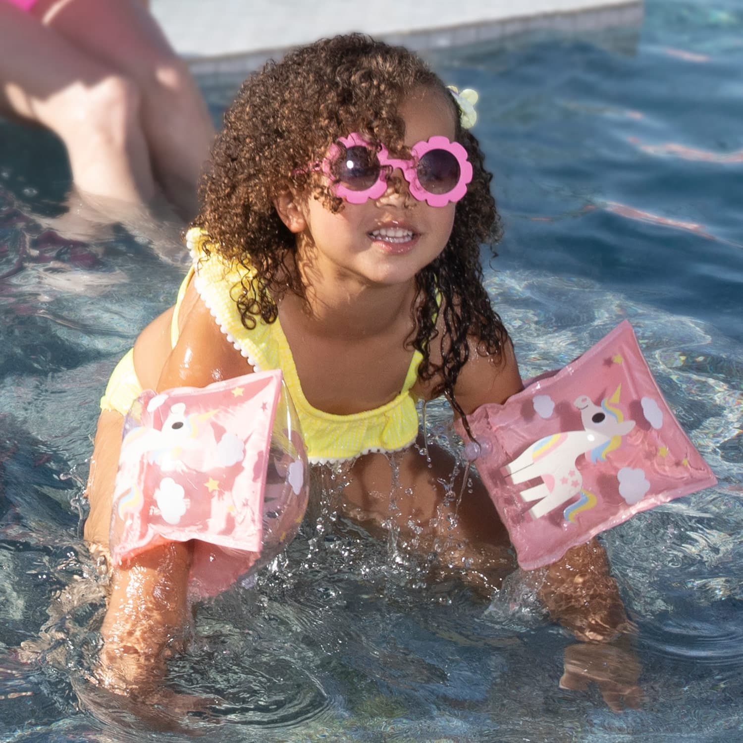 Child in a pool wearing pink sunglasses and arm floats with unicorn designs.