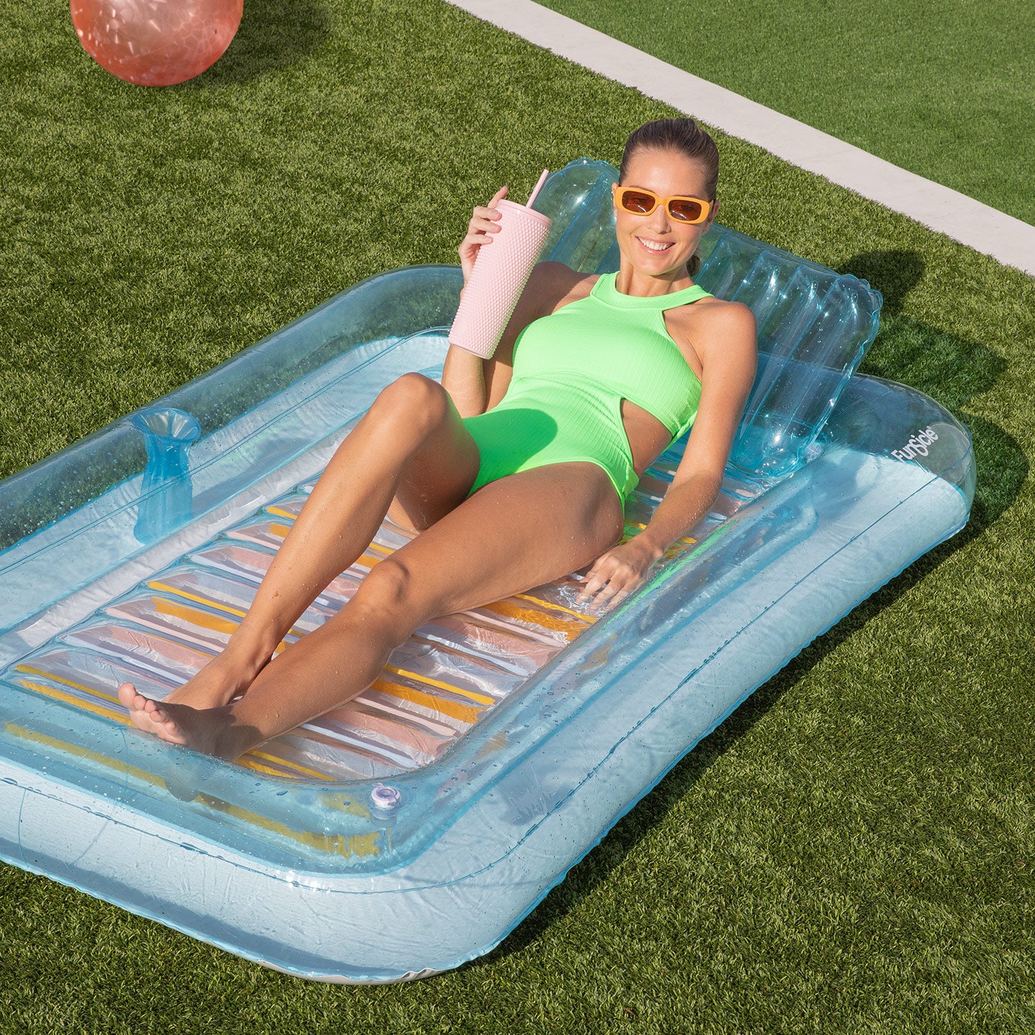 Woman in green swimsuit lying on a clear inflatable tub holding a pink cup