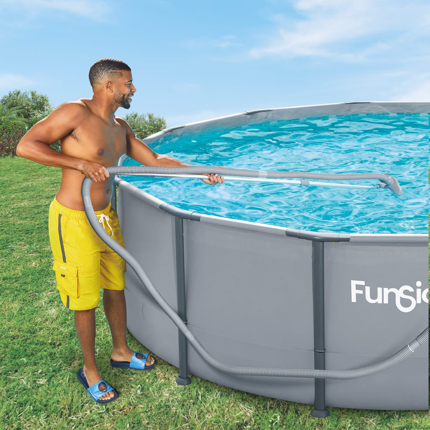 Man cleaning a above-ground pool with a hose, standing on grass with a clear blue sky.
